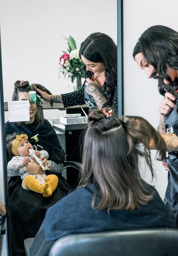 Woman sits in chair next to baby speaking with hair stylist